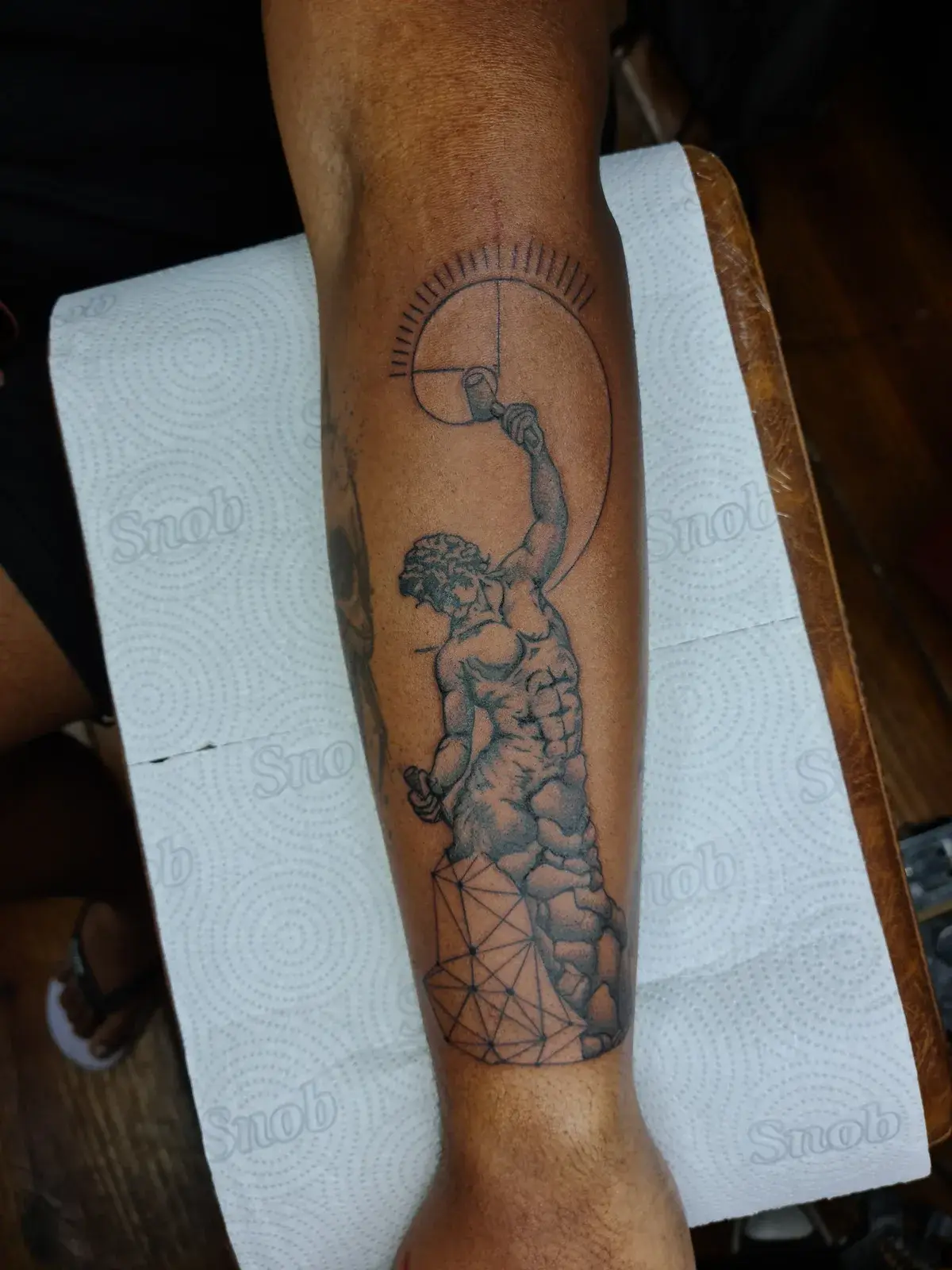 Tatuagem blackwork de figura clássica erguendo uma esfera, com geometria sagrada, no antebraço