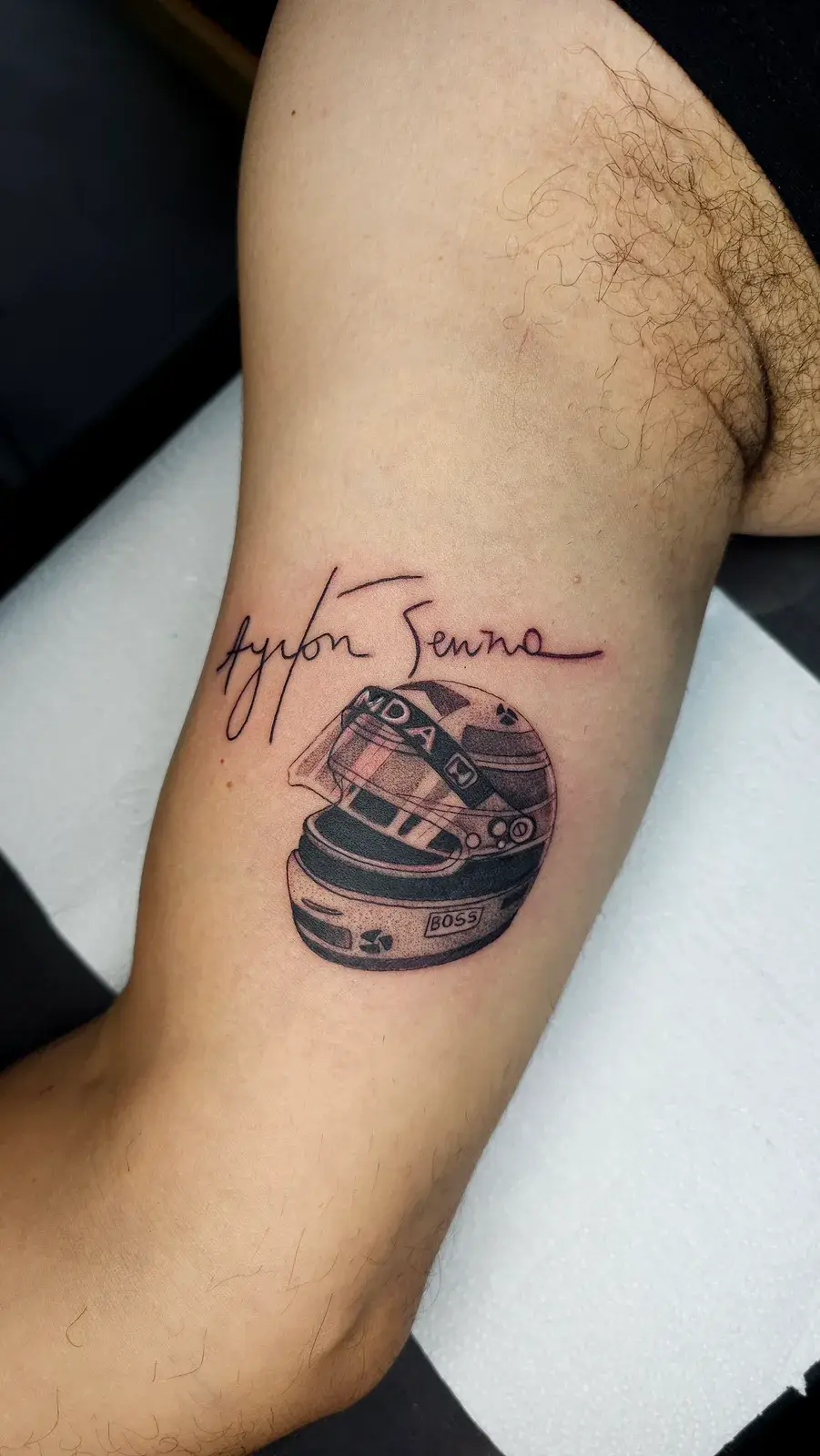 Tatuagem blackwork do capacete do Ayrton Senna com a assinatura dele, no braço, sobre o tríceps