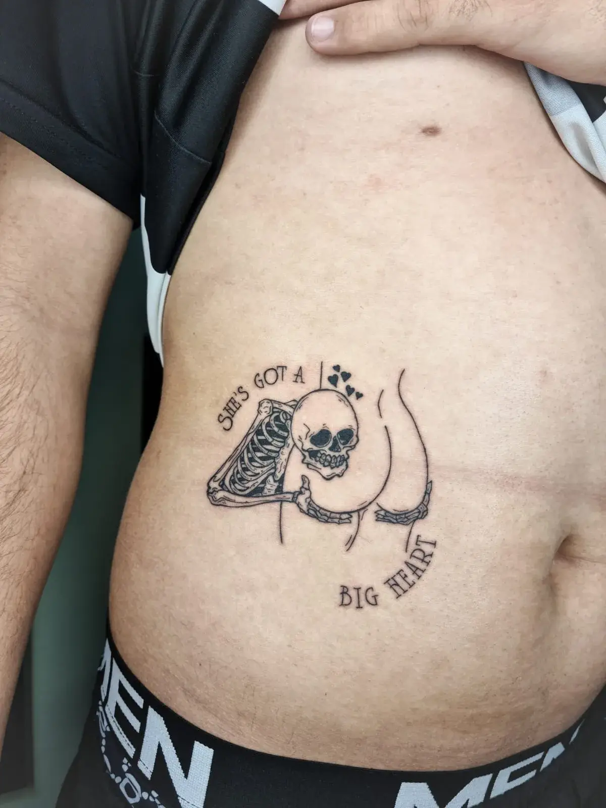 Tatuagem blackwork de esqueleto com a frase She's got a big heart, em traço fino no abdômen