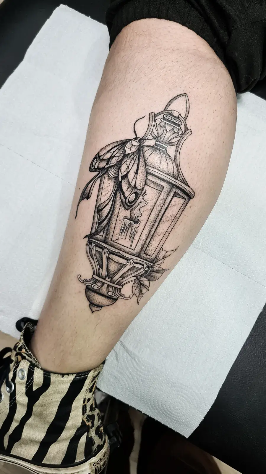 Tatuagem blackwork de lanterna antiga com mariposa e folhas, em pontilhismo na panturrilha