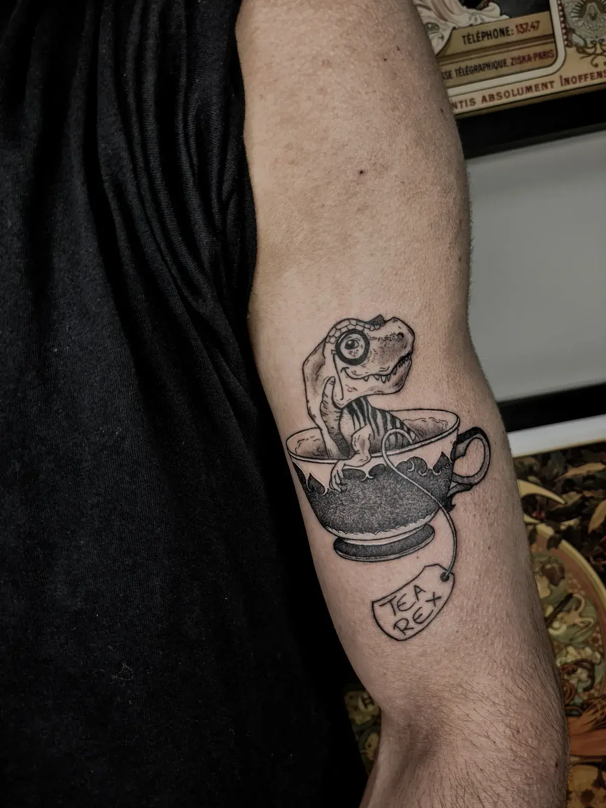 Tatuagem blackwork de T-Rex dentro de uma xícara de chá com a etiqueta Tea Rex, no braço, sobre o tríceps