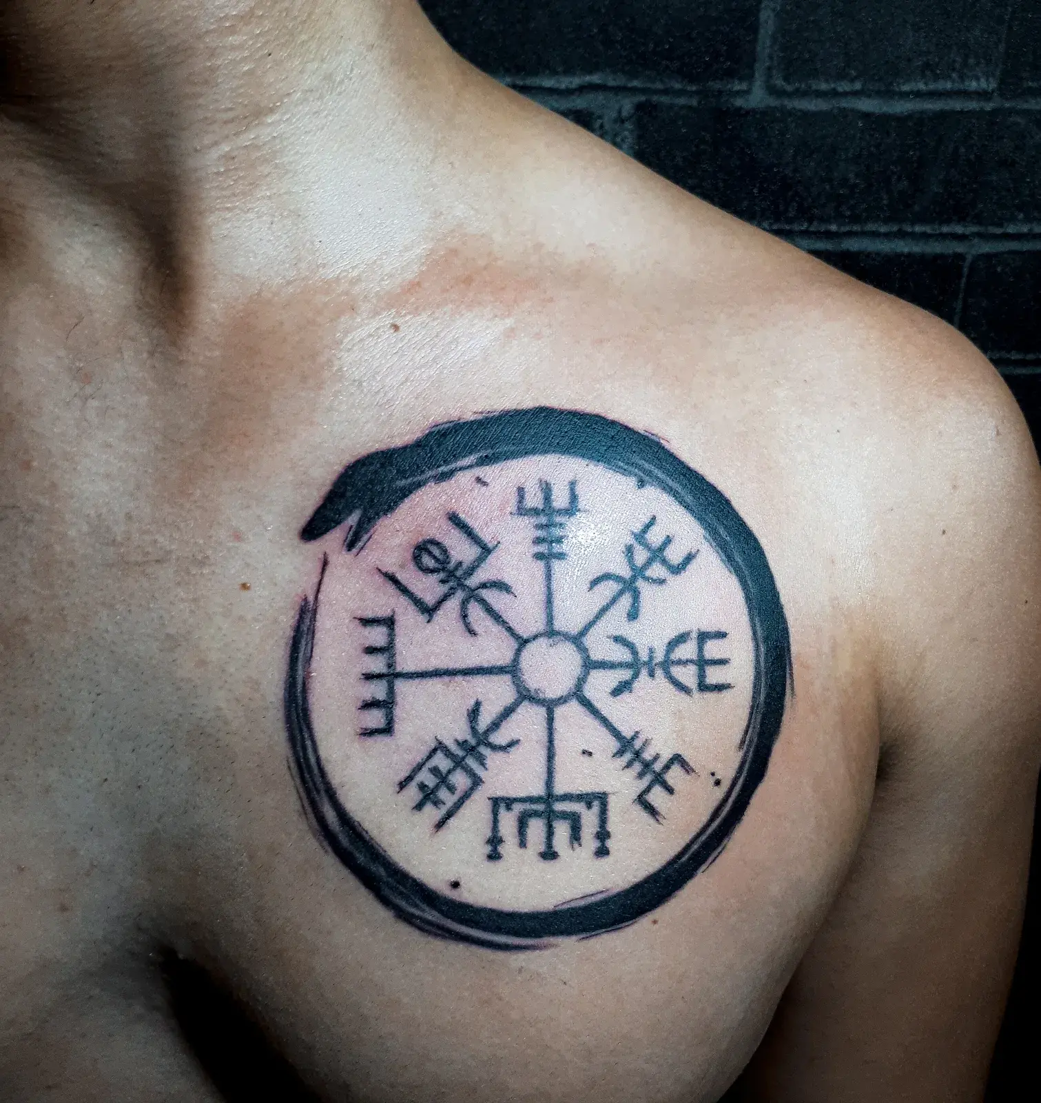 Tatuagem blackwork de uroboros envolvendo uma bússola nórdica, em preto sólido no peito