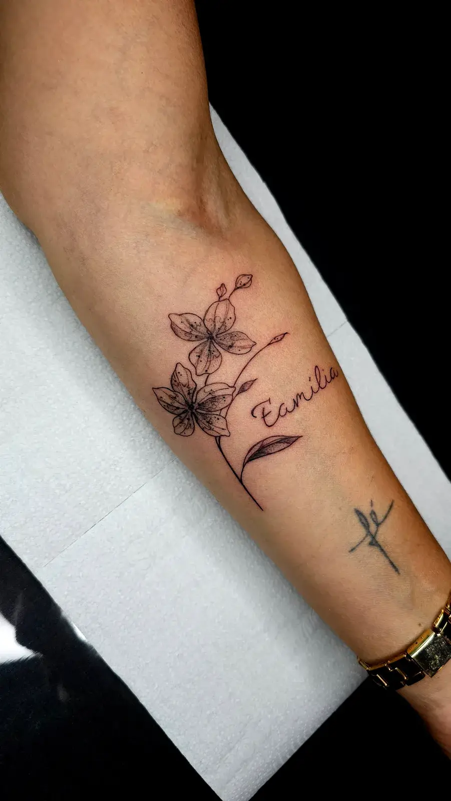 Tatuagem botânica com flores em traço fino e lettering no antebraço, vista de frente