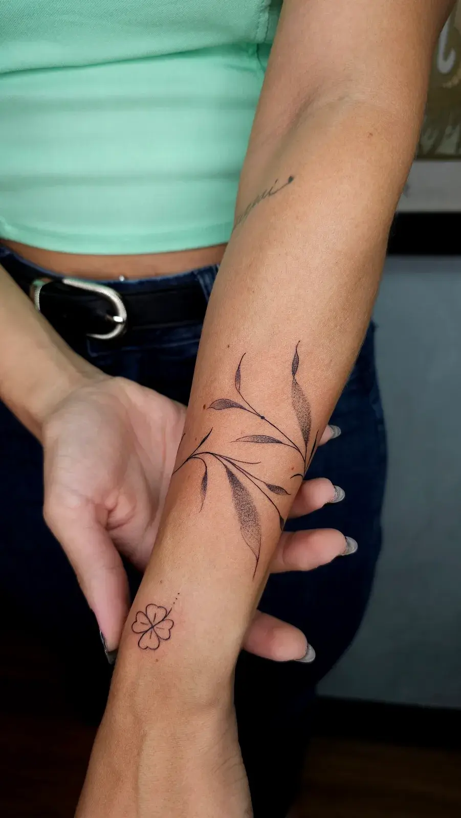 Tatuagem botânica de ramo com folhas contornando o antebraço, em traço fino com pontilhismo