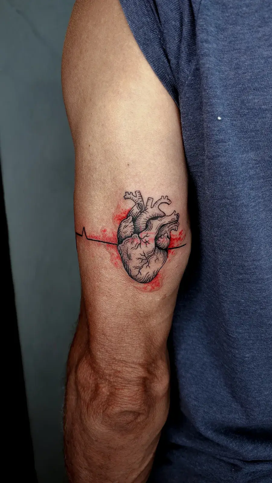 Cobertura de tatuagem com coração anatômico e linha de eletrocardiograma no braço, sobre o bíceps