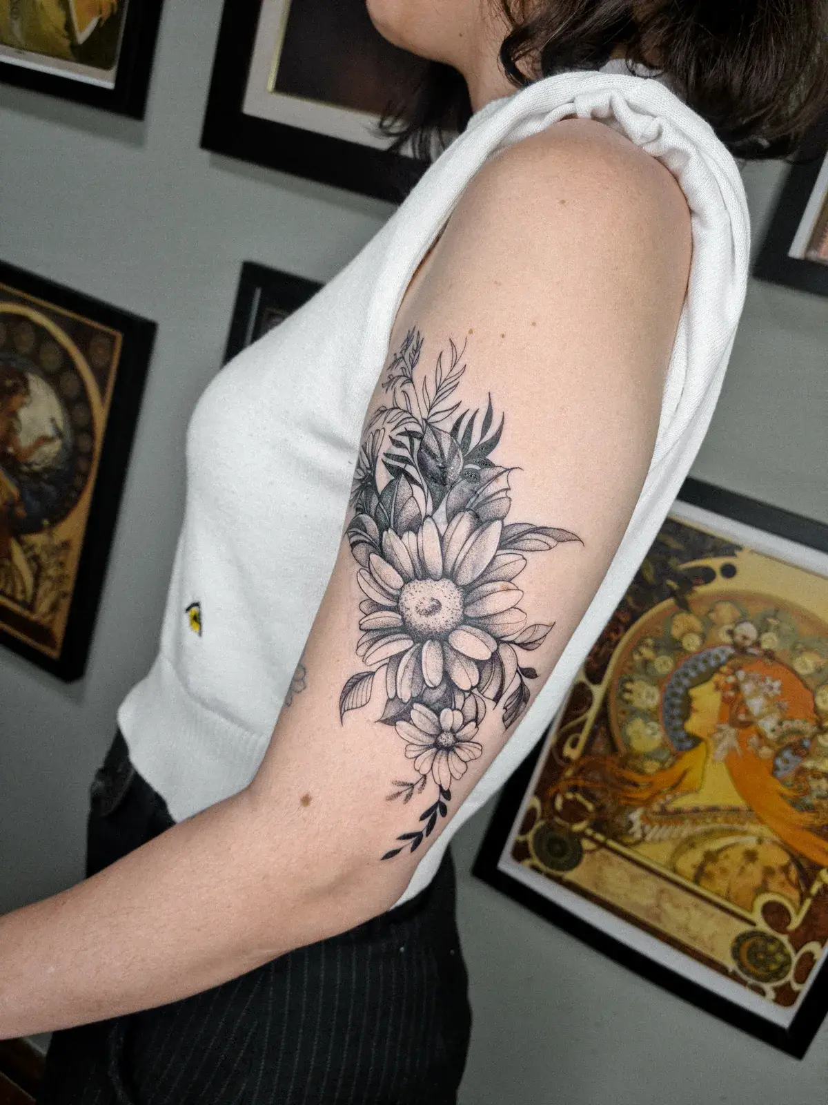 Cobertura de tatuagem com margaridas e folhagem em preto e cinza no braço, sobre o tríceps