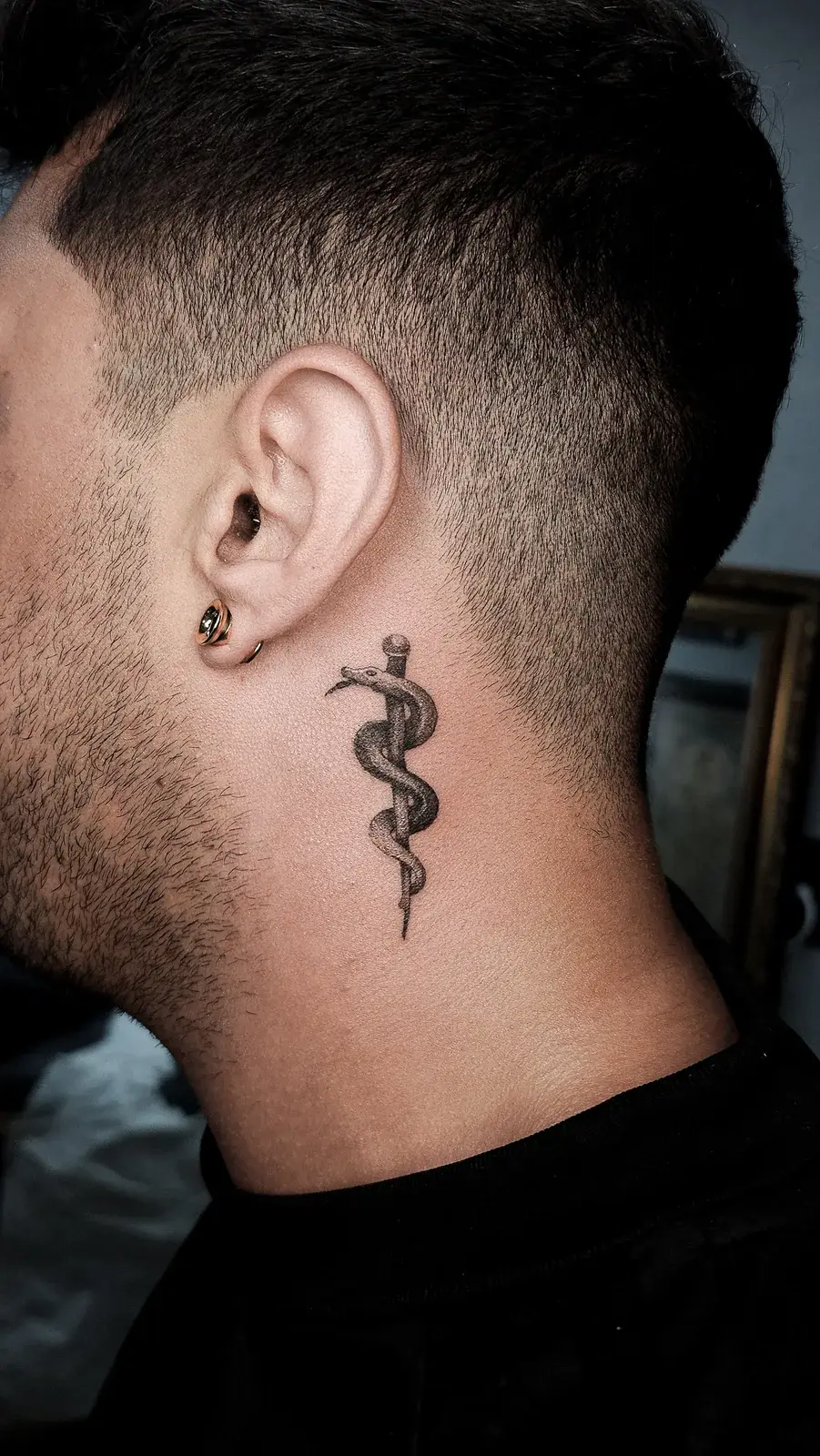 Tatuagem fine line do bastão de Asclépio em pontilhismo, atrás da orelha