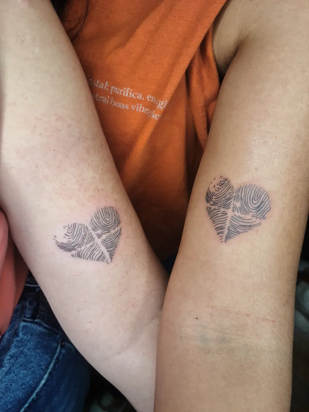 Tatuagem fine line de corações formados por digitais, no braço de duas pessoas