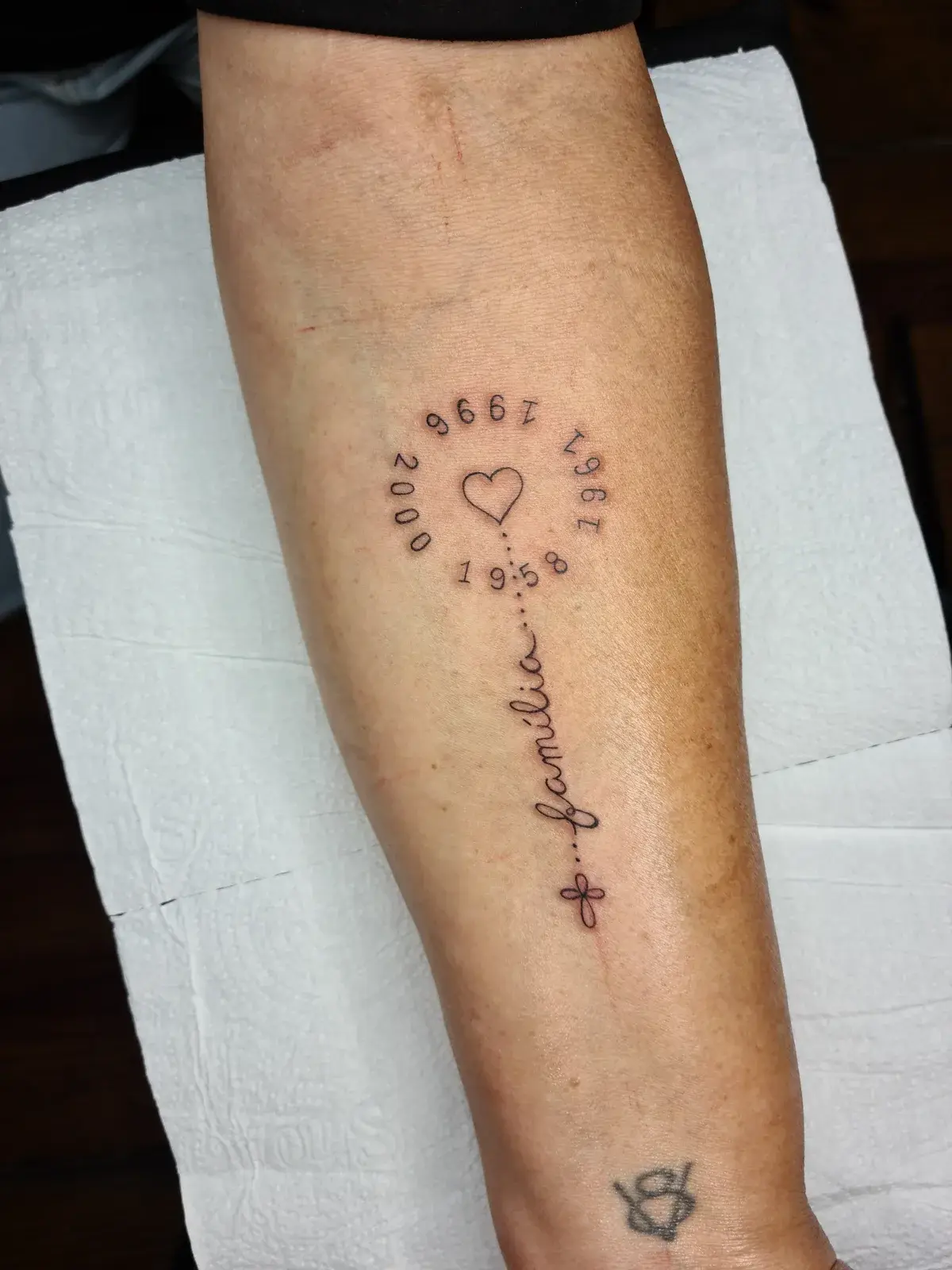 Tatuagem fine line com datas em círculo, coração, a palavra família e um terço, no antebraço