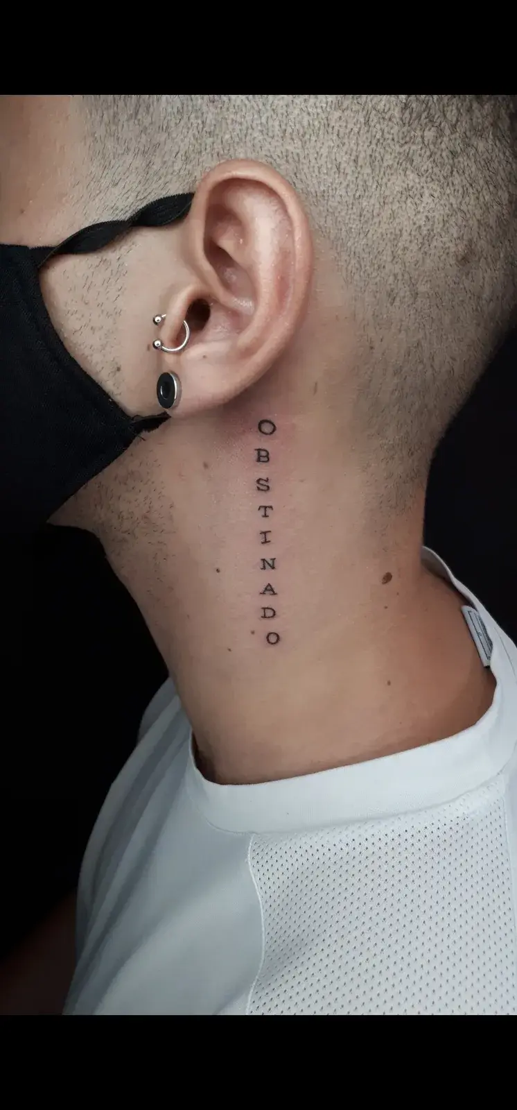 Tatuagem fine line com o lettering Obstinado na vertical, no pescoço