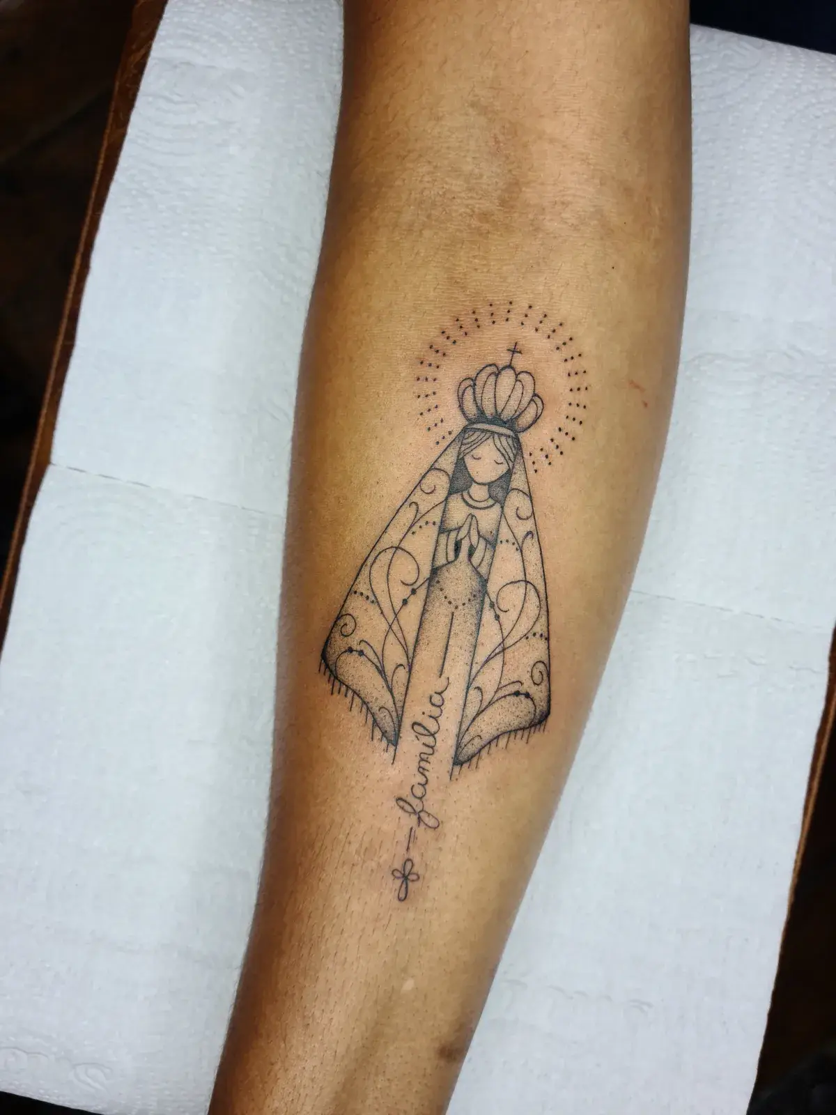 Tatuagem fine line de Nossa Senhora Aparecida com a palavra família e um terço, no antebraço