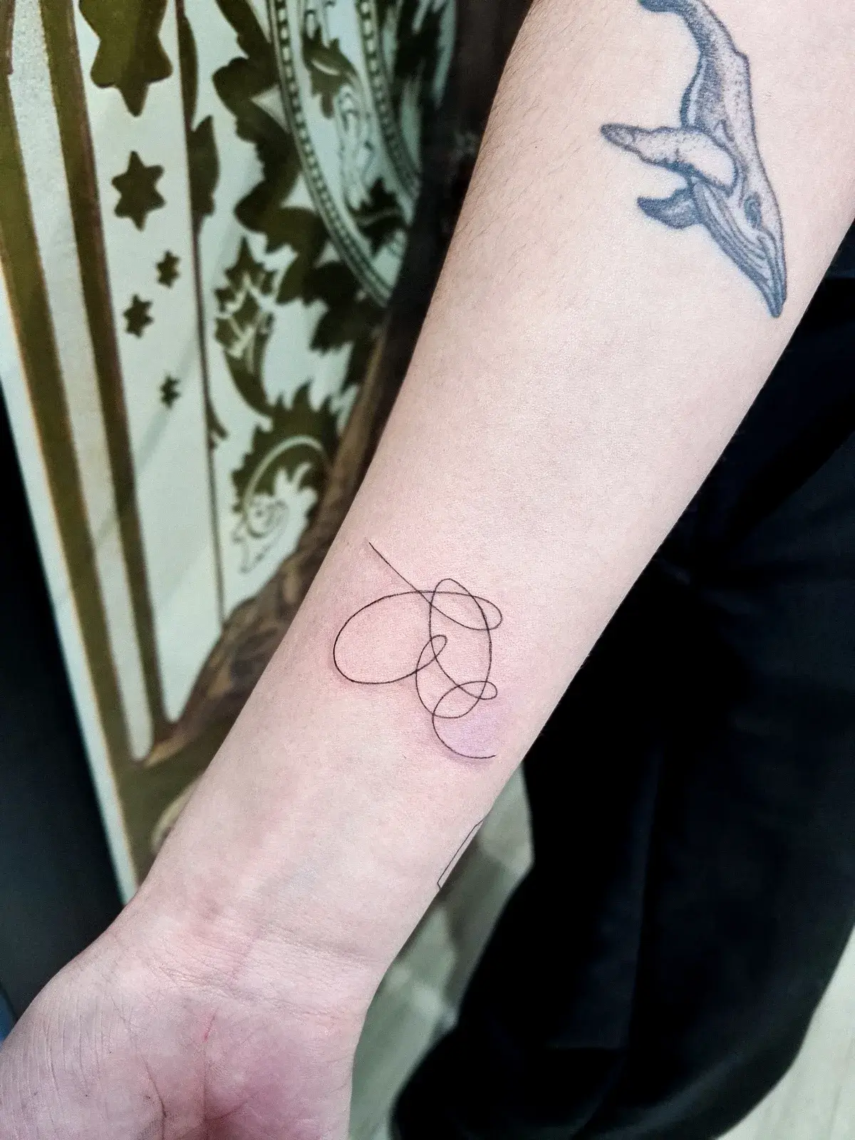 Tatuagem fine line com o símbolo do BTS em linha contínua no antebraço