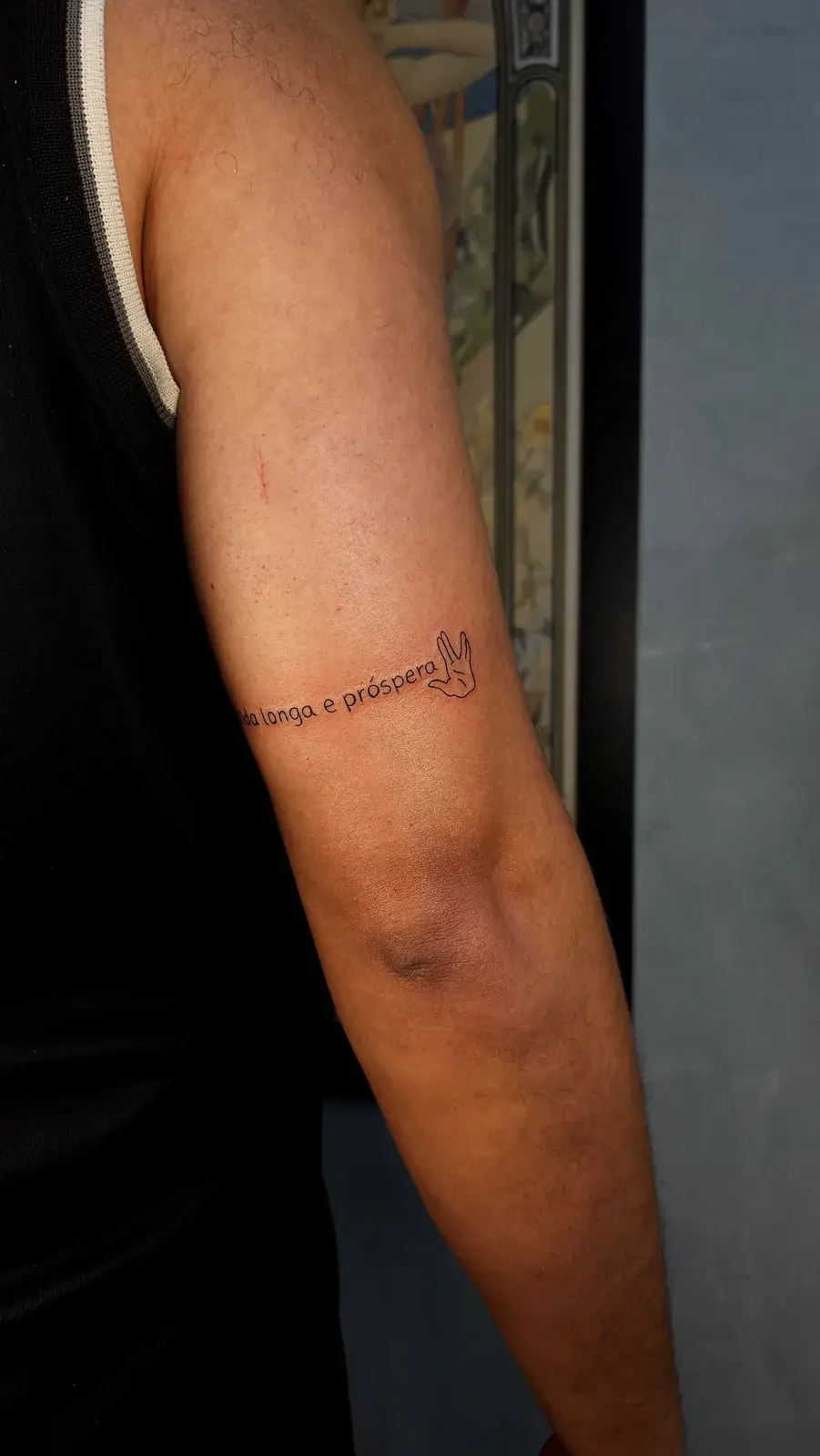Tatuagem fine line com a saudação vulcana de Star Trek e a frase Vida longa e próspera, no braço