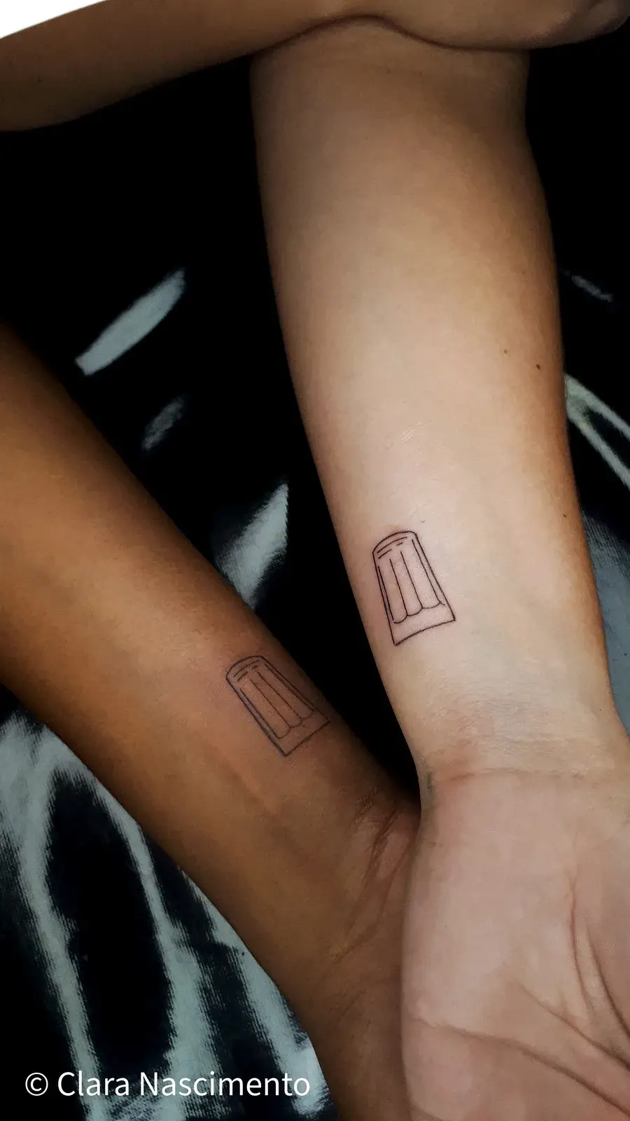 Tatuagem de amigos em fine line, com o copo da Lagoinha em traço mínimo no antebraço de duas pessoas