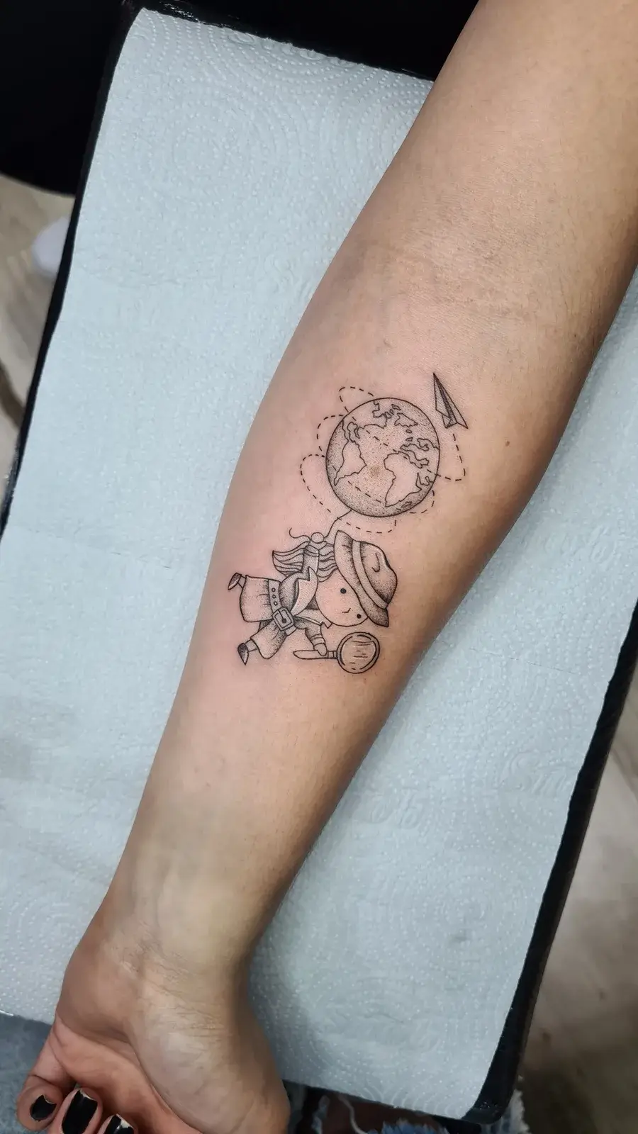 Tatuagem fine line delicada sobre viagens, com globo, avião e viajante de mochila, no antebraço