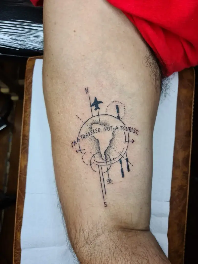 Tatuagem fine line com globo, bússola e a frase I am a traveler not a tourist, no braço