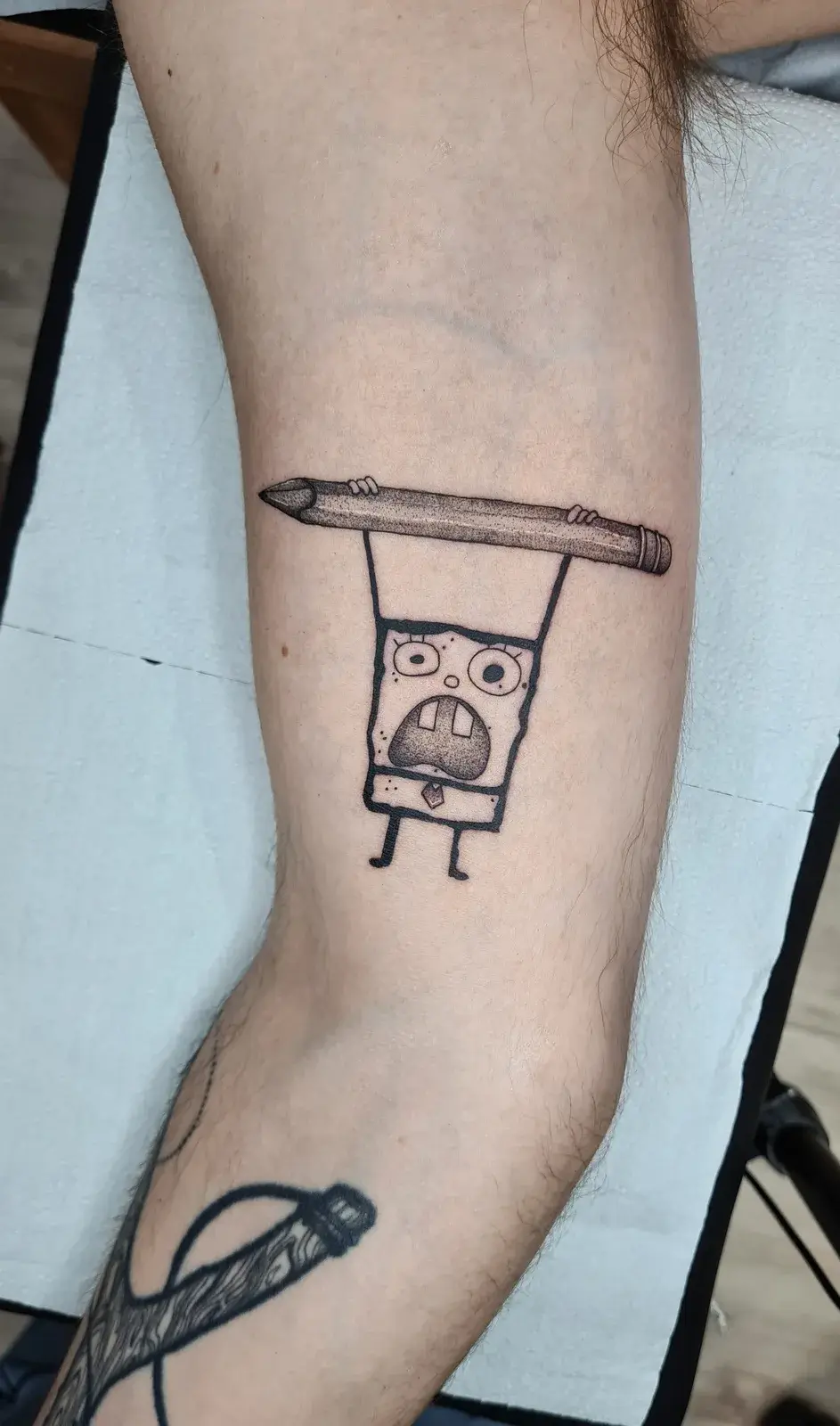 Tatuagem geek do Bob Esponja em traço de rabisco pendurado em um lápis, em blackwork no braço