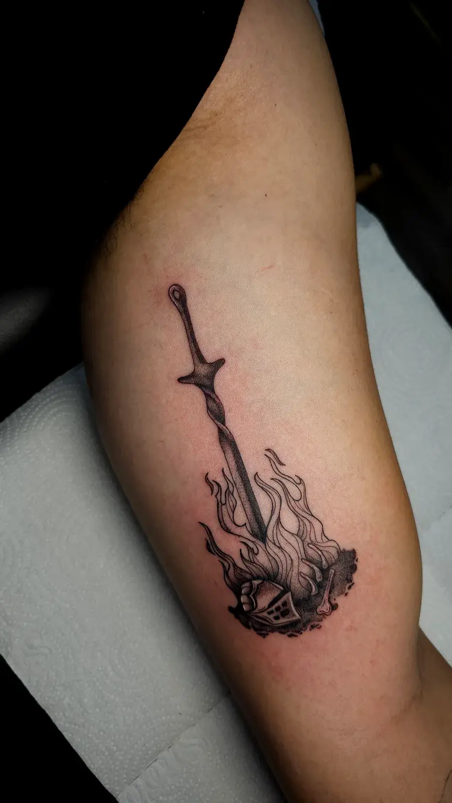 Tatuagem geek de Dark Souls com espada cravada na fogueira, em preto e cinza no braço