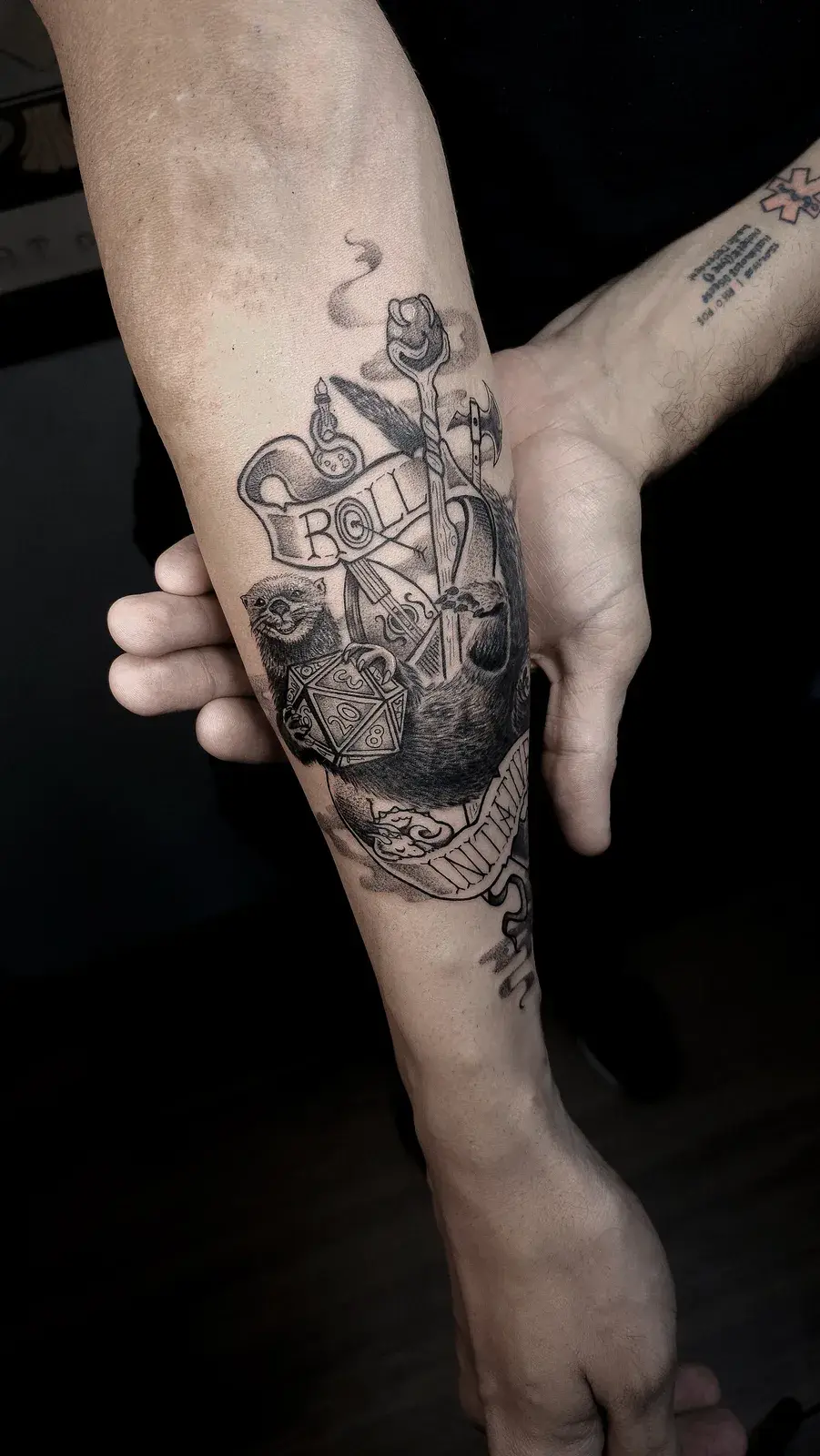 Tatuagem geek de Dungeons & Dragons com dado d20 e a fita Roll Initiative, em blackwork no antebraço