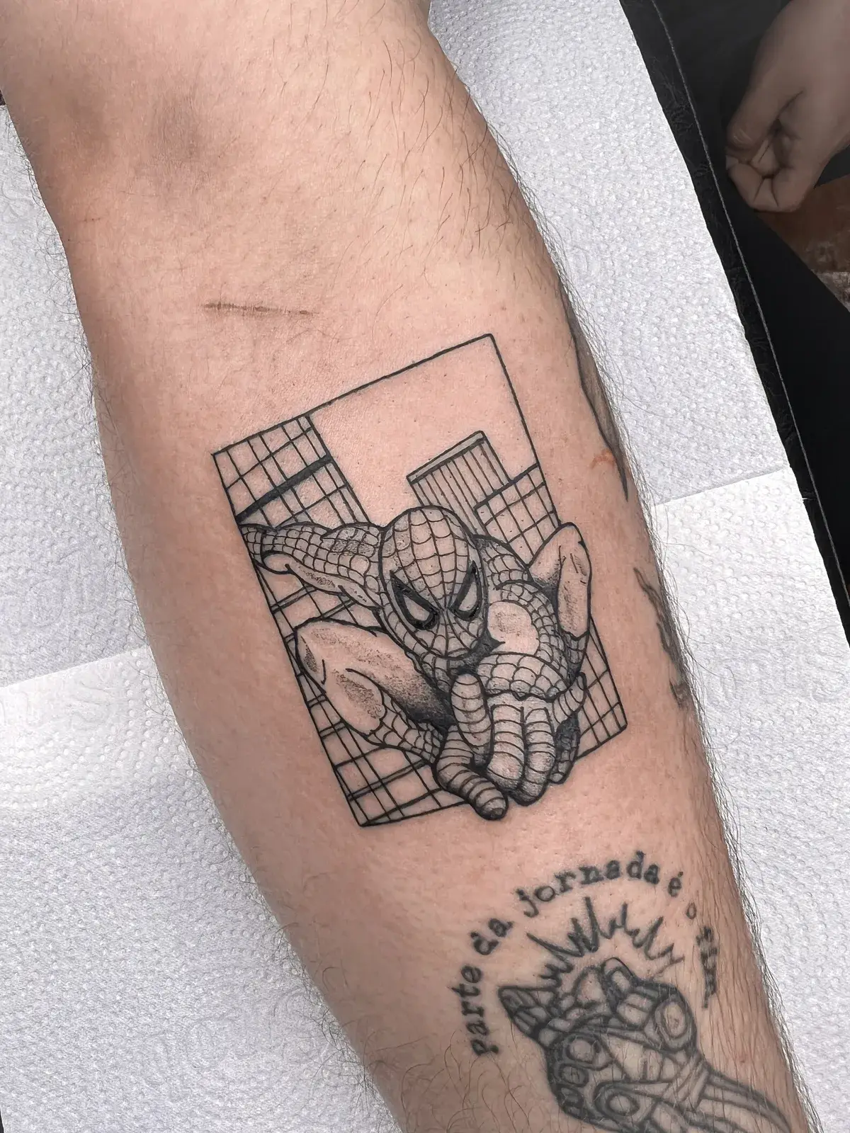 Tatuagem geek do Homem-Aranha entre prédios, em quadro com traço fino, no antebraço