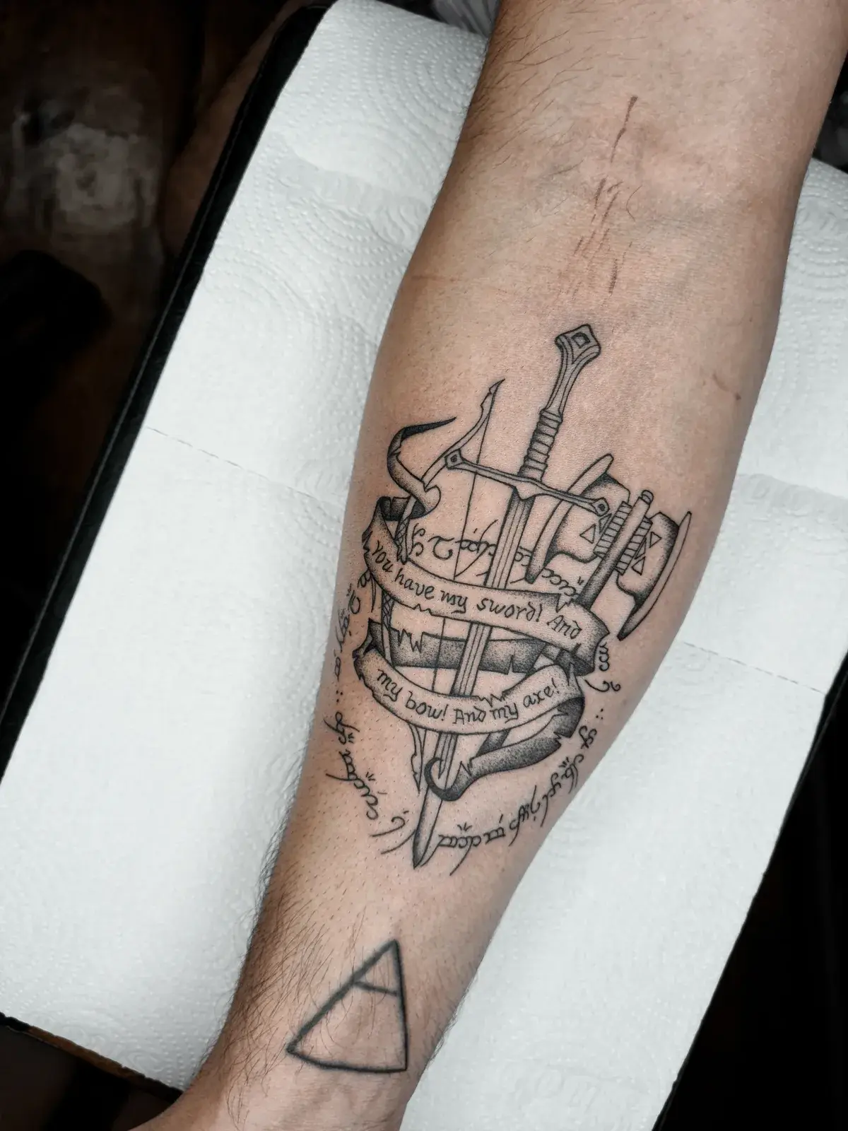 Tatuagem geek de O Senhor dos Anéis com espada, arco e machado no antebraço, vista pela lateral