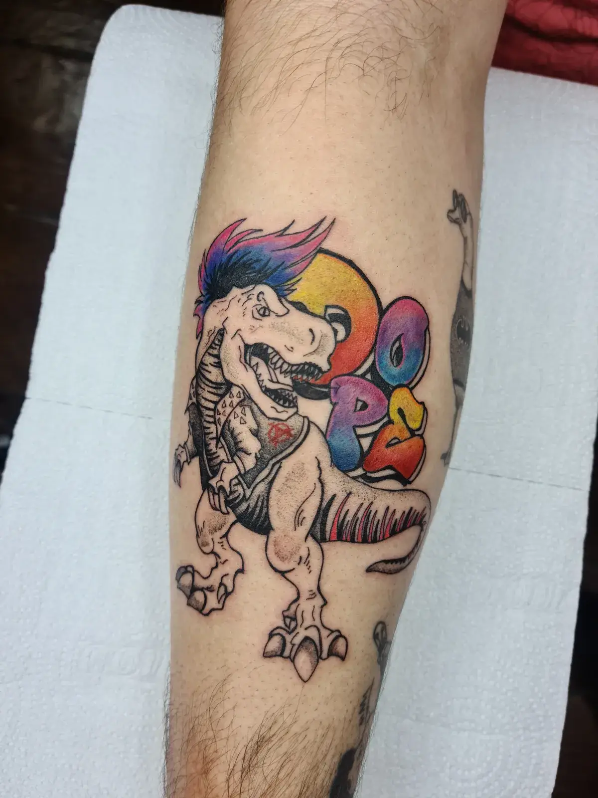 Tatuagem geek de T-Rex punk com moicano colorido e lettering em grafite, na perna