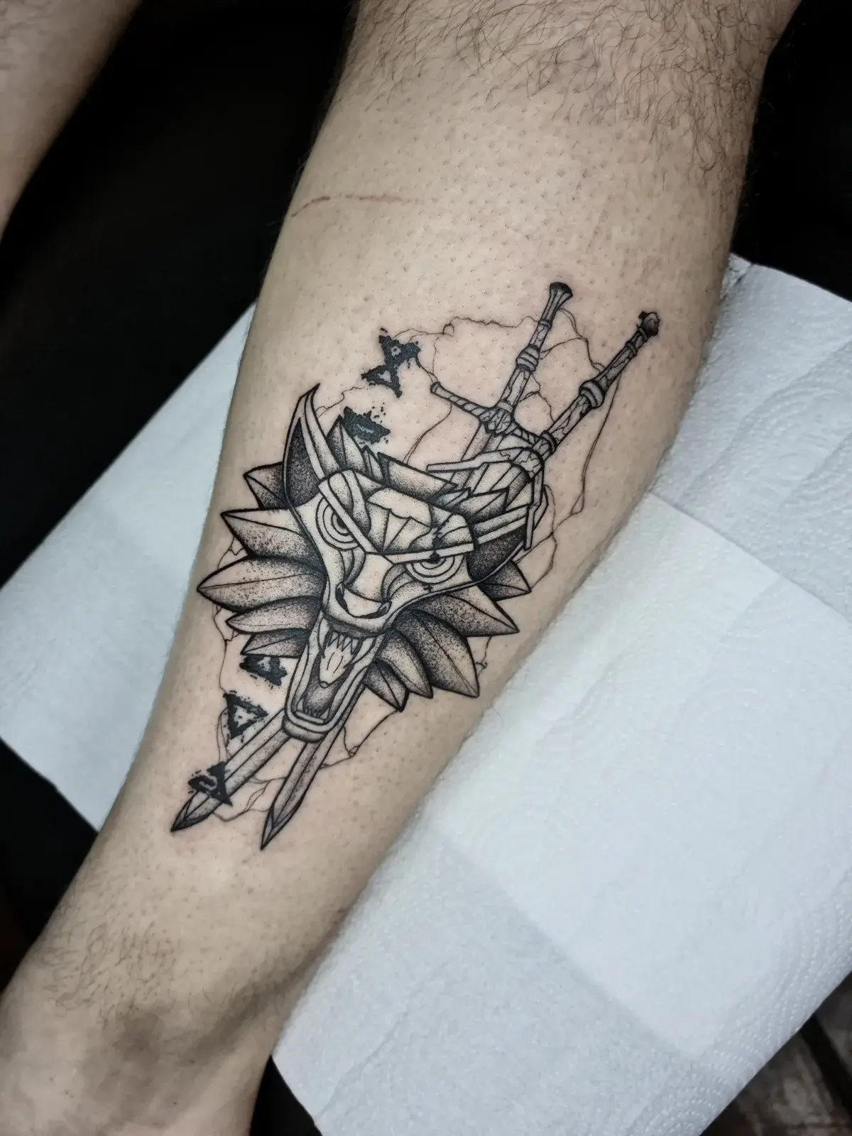 Tatuagem geek de The Witcher com o medalhão do lobo e as duas espadas, em blackwork na panturrilha